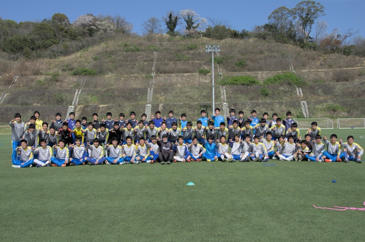 桜の咲くグラウンドで、大人数の部員と指導者が並んで記念撮影